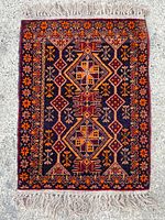 Full view of the small rectangular hand woven rug with geometric and floral pattern in red, orange, yellow, and black with fringes at edges.