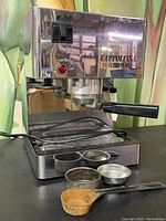 Front-right angled view of the Isomac Cappuccina espresso machine on a black surface showing portafilter, drip tray, switches, and included accessories in front (two filter baskets and scoop).