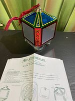 Hexagonal Mr. Christmas Magic Scene Ornament showing blue, green, red, and yellow stained glass effect panels with a red hanging ribbon and instruction sheet in front.