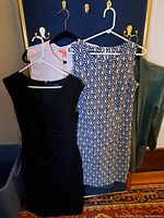 Photo showing three items of women's clothing hanging on hangers: a black dress, a black and white geometric patterned dress, and a pale pink long sleeve blouse.