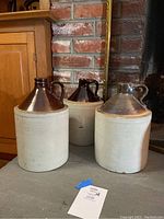 Three stoneware jugs with light beige bodies and various brown glazed conical tops, positioned on floor by brick fireplace and wood cabinet.