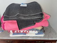Stack of blankets and quilts including a dark grey wool blanket and a bright pink fleece blanket on top of several folded quilts with various patterns.