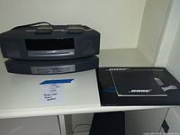 Two black Bose units stacked side by side with manuals on shelf