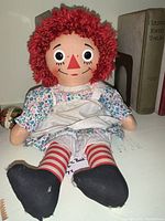 Raggedy Ann doll seated on shelf showing front details, floral dress, yarn hair, striped stockings, black shoes
