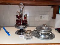 Photo showing silver plated cruette stand holding four cut glass bottles with red liquid and stoppers, and a small cut glass jar with metal lid placed in front.