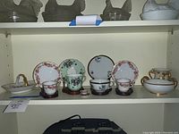 Shelf display of fine china collection including 4 tea cup and saucer sets with floral patterns, small decorative bowls, and a white vase with gold accents.