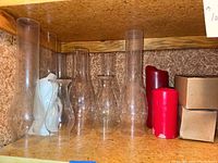 Photo showing nine clear glass hurricane chimneys of varying sizes and styles, plus four red candles, placed on a wood shelf inside a closet.