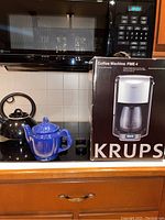 Krups coffee machine box with blue ceramic teapot and black tea kettle on kitchen stove.