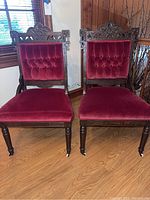 Front view of two antique wooden chairs with deep red velvet seat cushions and tufted backs, showing castored front legs and intricate wood etching