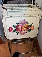 Six vintage metal TV trays with floral designs stacked on a metal frame with gold decorative accents.