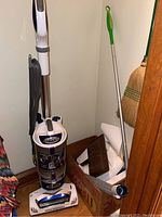 Photo showing Shark upright vacuum cleaner with cord, clear canister, and labels, positioned in corner beside broom and a box.