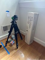 Photo of Slik U212 Universal Tripod standing next to its original box on hardwood floor. Shows the tripod extended with adjustable legs and mounting head with green bubble level.
