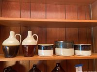 Two tapering stoneware jugs with tall necks and handles, with light colored glaze and brown lower halves, along with three cylindrical crocks with dark upper glaze and light lower glaze displayed on a wooden shelf.