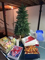 Full view of a green artificial Christmas tree in a gold pot on a table with open boxes containing various decoration items including colorful balls and ribbons.