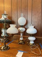 Three lamps on a wooden surface against a wood-paneled wall: two hurricane lamps with glass globes and one desk lamp with a corrugated white glass shade. The tallest lamp features hand-painted flowers on its white glass body.