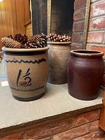 Three stoneware crocks of varying colors and shapes placed together with pine cones inside some crocks on a stone hearth in front of a brick fireplace.