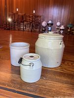 Group photo of three stoneware crocks including a large crock with lid, a medium crock with lid, and a cylindrical crock without lid, placed on wooden floor.