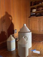 Three cream-colored stoneware jugs arranged on wooden floor against wood-paneled wall, showing varied sizes and forms, largest with number 5 marking.