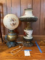 Two antique brass and glass lamps displayed side by side on a wooden surface with wood paneling background. The taller lamp has white glass with red floral decorations and ornate brass detailing including lions heads. The shorter lamp has a round white glass globe with floral patterns and a brass and cast metal base. Both lamps have glass chimneys and attached cords.