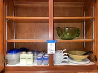 Full view of upper cabinet showing vintage kitchen items including green measuring bowl, salt and pepper shakers, white crocks, small juice glasses, and plastic containers.