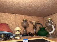 Five lamps on a wooden shelf, featuring two clear glass hurricane lamps, one green glass lamp, one beige ceramic lamp, and one brass lamp with red fabric shade.