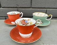 Three antique Aynsley cups and saucers showing distinct color and floral patterns, gold trim, and good condition.
