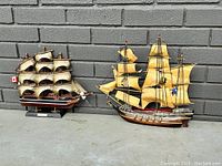 Two model clipper ships shown side by side on the ground against a gray brick wall, showcasing their sails and details.