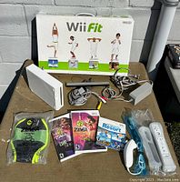 Nintendo Wii console and accessories including sensor bar, power adapter, two Wii remotes, Nunchuk controller, Wii Fit balance board, and four Wii game cases arranged on a table outdoors.