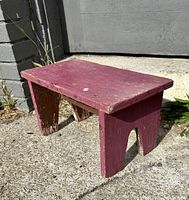 Side view of small wooden stool painted dark red with chipping paint and weathering.