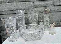 Full lot showing all six cut crystal pieces arranged on table against stone wall: includes three tall vases each with distinct cut patterns, footed bowl, decanter, and condiment jar.