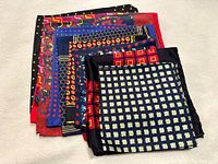 Stack of multiple silk pocket squares showing colorful geometric patterns predominantly in black, blue, red, and yellow