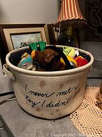 Canvas basket with text 'I never met a toy I didn't bite' filled with assorted dog toys placed on a table with lamp and framed picture in background.