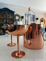 Photo showing one copper champagne glass and copper ice bucket with reflections and background furniture