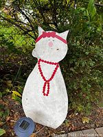 Photo of a flat wooden cat figure painted white with red bow and red beaded necklace around its neck, placed outdoors on grass near plants and a chain link fence.