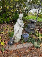 Image shows a concrete garden statue of a woman draped in cloth, standing on a base outdoors with surrounding garden elements including two black solar garden lights and a decorative urn.