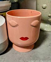 Photo of a pink ceramic flower pot shaped like a face with painted closed eyes and red lips, standing on a pedestal base.