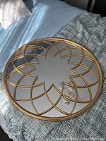 Round metal mirror resting on a blue bedspread with a gray knitted blanket nearby, showing clear reflective surface and decorative gold metal frame with petal-like segmented design.