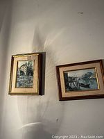 Photo of two vintage framed prints hung on a wall, one depicting a watermill by a river and the other a dock with boats on a rocky coast.