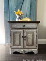 Front view of the vintage nightstand showing distressed white and blue paint, black hardware, and stained wood top with decorative objects.