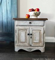 Full view of vintage nightstand showing distressed white and blue painted finish with stained wood top. One drawer and double doors with black knobs and decorative shell carving at base.