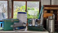 Wide angle photo showing a variety of camping items: large green Coleman propane stove, Igloo drink cooler, metal thermos, stove accessories, and other items on table by window.