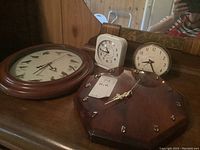 Four clocks on wooden surface: large wall clock with bird silhouettes, octagonal wooden clock with gold numerals and hands, Westclox Big Ben alarm clock, and smaller white cubical clock.