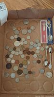 Overview of assorted coins spread on brown paper alongside three collectible spoons, one in a case.