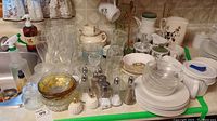 Wide view showing varied glassware, ceramic plates, bowls, salt and pepper shakers, milk jugs, tea set components arranged on table.