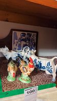 Grouped view of two rooster figurines, two white ceramic cow figurines, and small service tray on patterned surface under wooden shelf