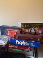 Full view of stacked board games and puzzle book showing Rummoli tray, People trivia game, Entertainment game, a crossword puzzle book, and a domino set in brown case.