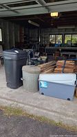 Photo showing black resin waste bin on wheels, vintage aluminum trash can, and gray plastic storage bin with white lid in garage setting.