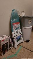 Photo shows gray plastic wash bucket, white folding step stool, countertop ironing board wrapped in plastic, electric iron and box for Rubbermaid Ironing Organizer also visible.