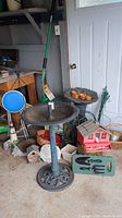 Full overview photo showing large dark green plastic pedestal bird bath, smaller grey bird bath, pink house bird feeder, hand tools on knee mat, garden ornaments, wicker baskets, garden grabber rake, and small folding stool clustered near a garage door.