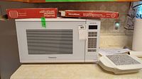 Panasonic microwave NN SG636W with control panel and digital screen, placed on countertop with boxed cooking sheet above.
